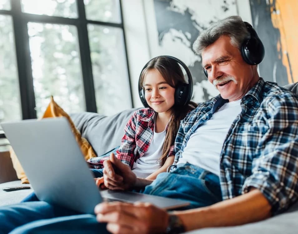 Avô e neta no sofá de casa, de fones de ouvido, assistindo a algo juntos no notebook